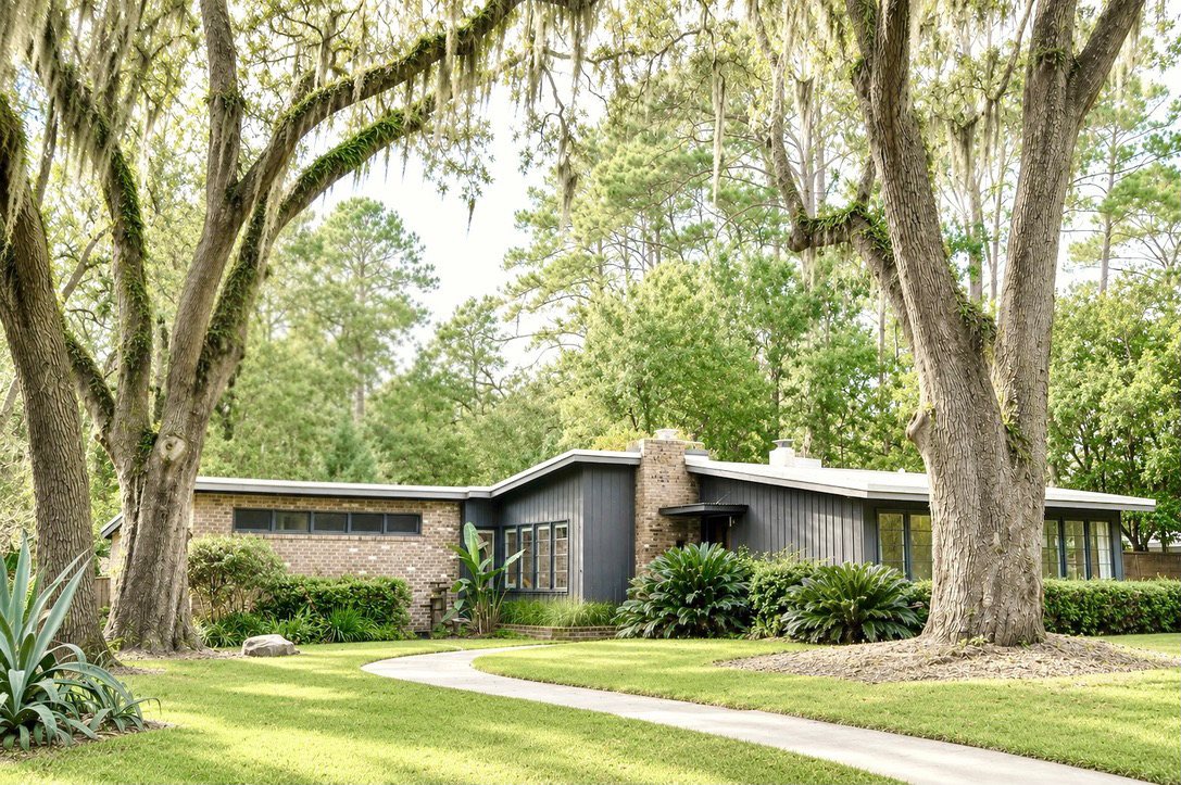 Front exterior of the mid-century modern home beneath moss-draped live oaks, with a curved walkway across the lawn